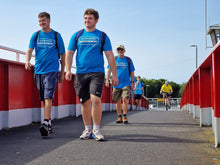 Load image into Gallery viewer, Two men in blue shirts walking on a bridge with a clear blue sky.